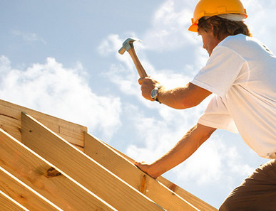 Dachdecker mit gelbem Schutzhelm beim Befestigen von Konstruktionsvollholz (KVH) auf einem Dachstuhl unter strahlendem Himmel.